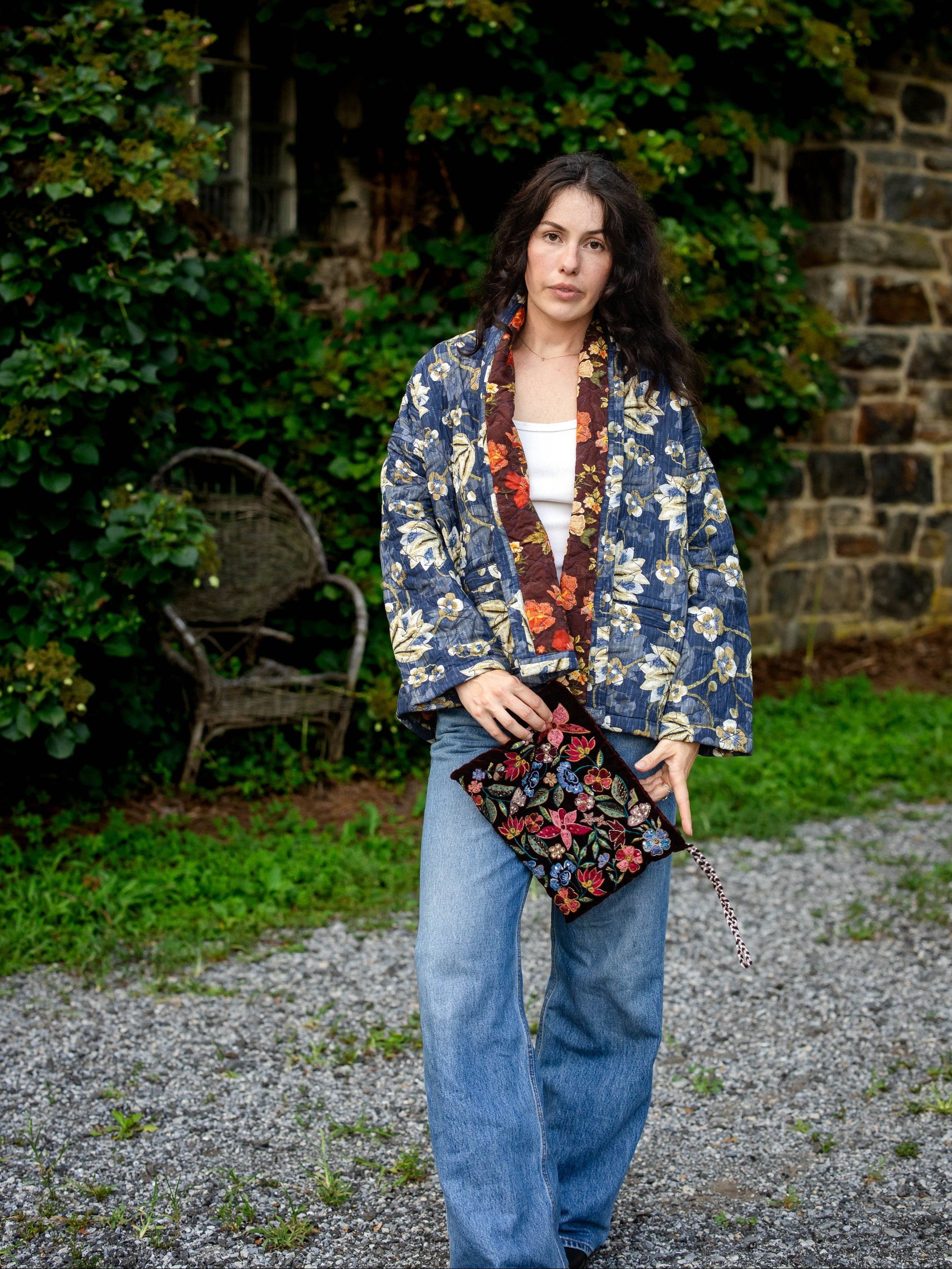 Woman in a floral jacket and jeans holding a clutch in front of a stone wall and greenery