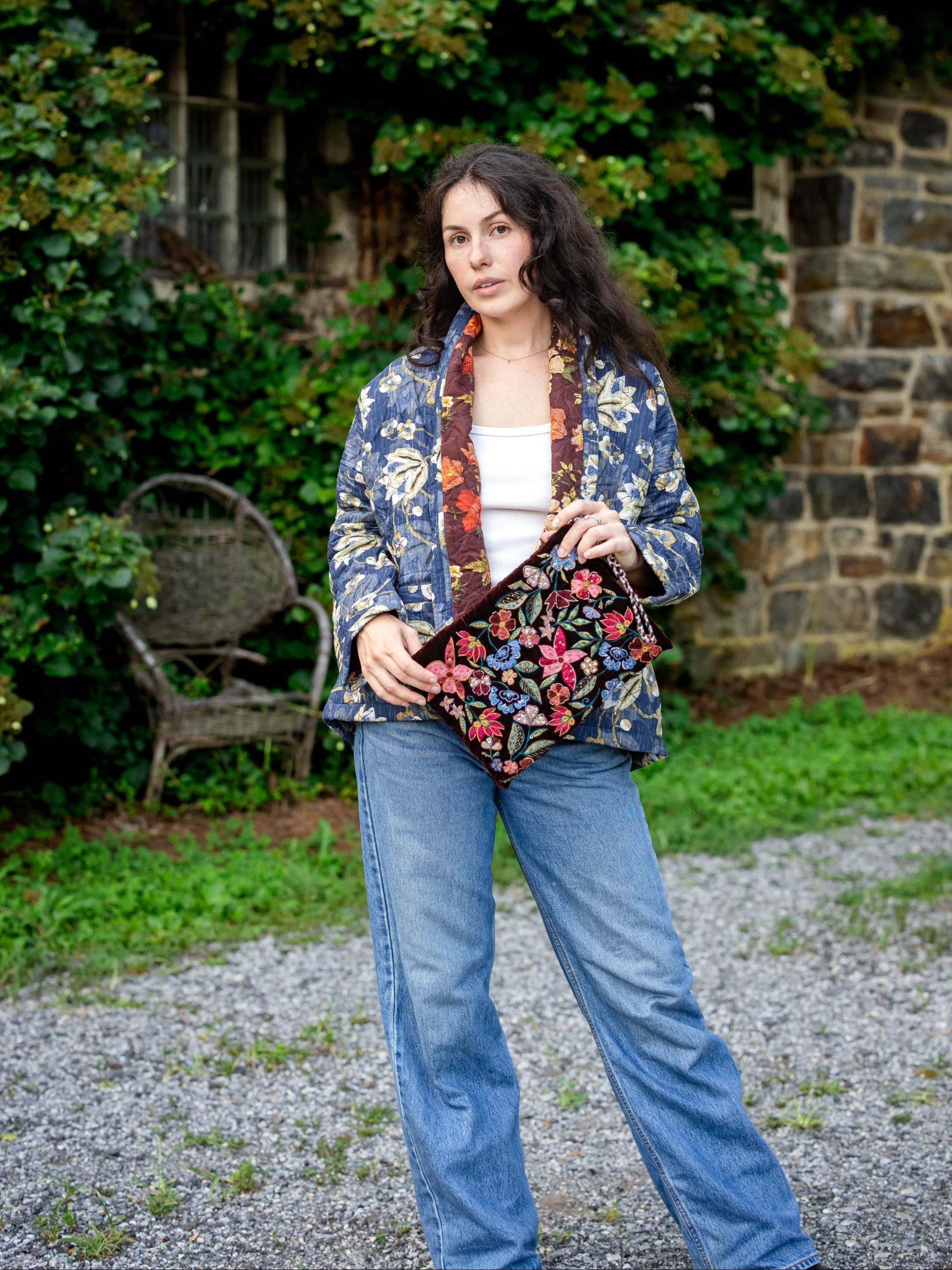 Woman holding a floral clutch in front of a stone wall and greenery